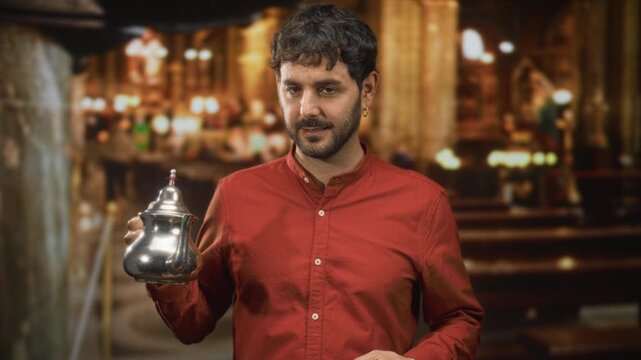 Man with beard and earring holds a silver teapot in his hand, presenting and examining the kettle while standing amid wooden pews in a building; quiet contemplation.