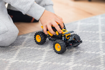 Close-up of a child's hand playing with a yellow toy monster truck on a grey carpet. Concept of early childhood development, creative play, and motor skills at home. © Sheviakova
