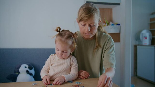 Mother and toddler sit at a table, focused on assembling a puzzle together. The toddler, with pigtails, concentrates intently on the pieces. The mother guides gently, her expression warm and patient