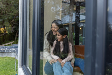 Indian mother and daughter sitting on cushioned seat, holding, looking through dark window at yard © wavebreak3