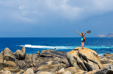 Carefree woman standing on huge rock with arms raised holding hat looking at the Atlantic ocean in Galicia, Spain. Concept of Freedom, success and achievement © M.studio