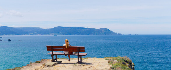 Panoramic view of woman sitting on a wooden bench on a cliff overlooking the Atlantic ocean, peaceful and tranquil scene © M.studio