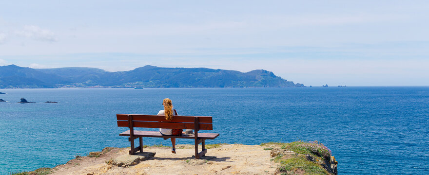 Panoramic view of woman sitting on a wooden bench on a cliff overlooking the Atlantic ocean, peaceful and tranquil scene