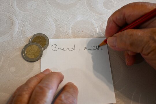 Close-up of an elderly woman's hand writing a shopping list next to a few euro coins, representing budgeting, cost of living, and daily life, Copy Space for text.