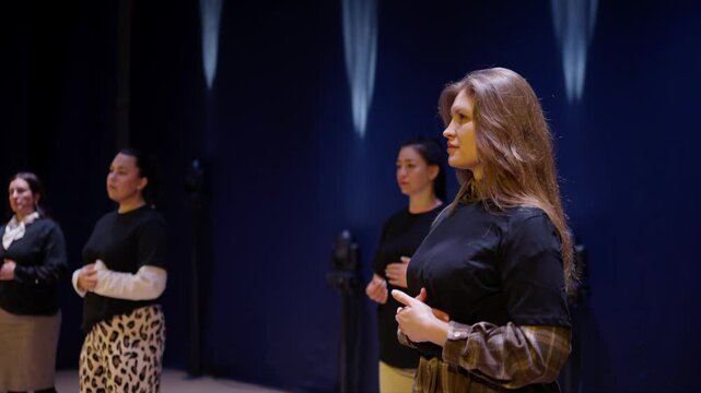Four women standing in a row on a dimly lit stage, rehearsing a performance with synchronized hand gestures against a dark blue theatrical background