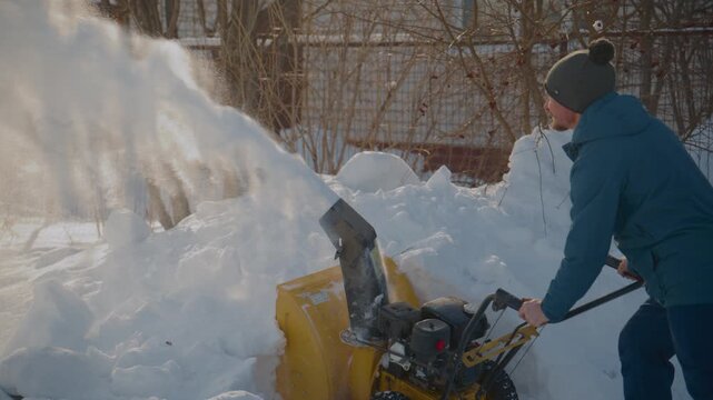 Person pushes snow blower across snowy yard. Machine throws snow high into air. Sunlight glares off snow, creating bright reflections. Person wears winter hat and coat, focused on task