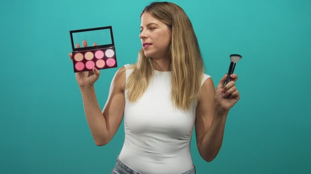 Woman holding pink blush palette and kabuki brush, eyes closed while lifting brush to cheek against teal backdrop in studio; confidence focus.