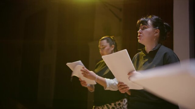 Focused performers rehearsing a dramatic script on a dimly lit stage, reading from printed pages under warm theatrical lighting during an intimate acting practice