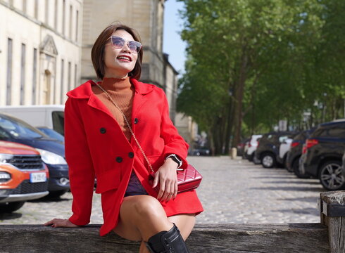 A woman wearing a vibrant red coat and sunglasses sits on a wooden beam along a city street with parked cars and classic architecture in the background