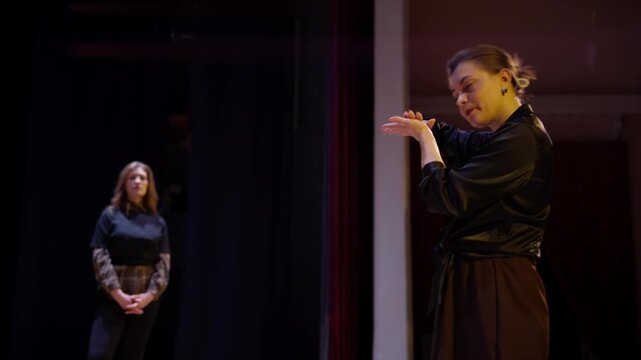 Interpreter uses expressive sign language to communicate with performers during inclusive theater rehearsal on dimly lit stage with dramatic spotlighting