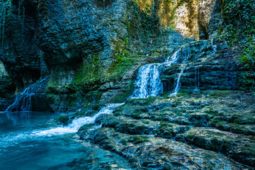 A small waterfall cascades over mossy limestone ledges into the turquoise water of Martvili Canyon, with steep rock walls and dense greenery framing the scene. © Auggie