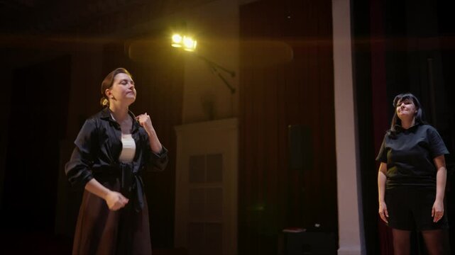 Passionate woman rehearsing dramatic monologue on dimly lit theater stage with attentive partner during acting workshop under warm spotlight