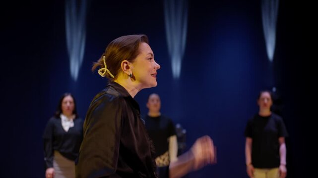 Passionate woman delivering dramatic monologue on dimly lit stage with focused spotlight as attentive performers stand in background during rehearsal