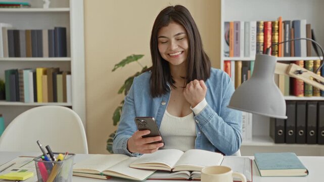 Woman holding smartphone and open books, hand near chest and smiling while seated at a desk in a building; study focus contentment.