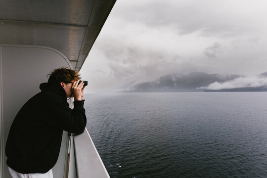 Teen boy looking through binoculars on cruise balcony at ocean
