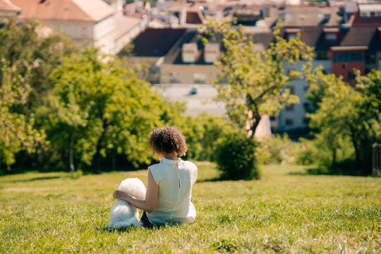 Woman sits on grass with Bichon Frise dog in Prague park sunny day