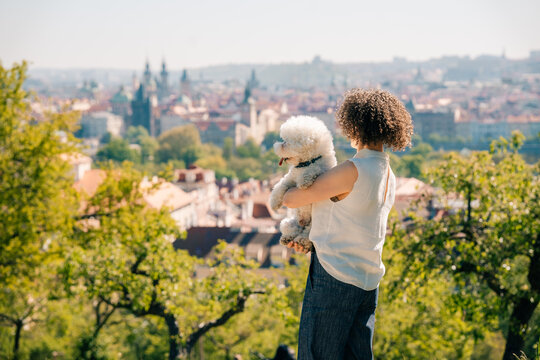 Woman holds Bichon Frise dog while looking at Prague from hill