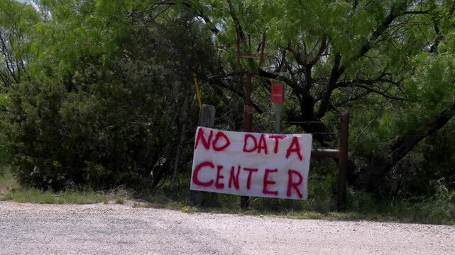 A homemade white sign, boldly painted with 'No Data Center' in red, stands against a backdrop of lush green trees. It signifies local community resistance and environmental concerns regarding proposed