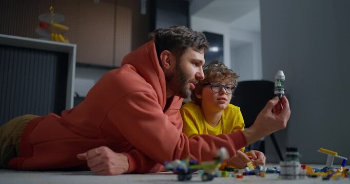 Father and young son lying on the floor building a toy rocket together, sharing a focused creative moment with colorful construction pieces at home
