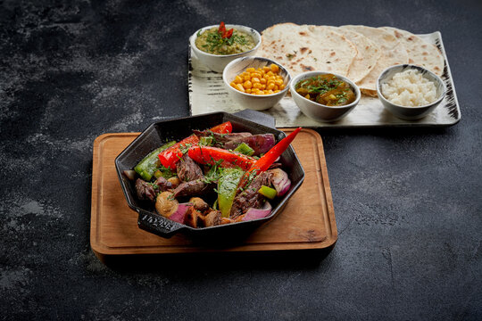 Traditional Mexican beef fajitas in a cast iron skillet served with tortillas, guacamole, corn, and rice. High-end ethnic food photography on a dark stone background.