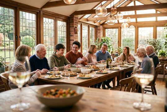 A multi-generational Caucasian family gathers for a home-cooked meal around a long wooden dining table in a sunlit, wood-accented sunroom.