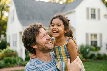 Fototapeta premium A smiling father holds his joyful mixed-race daughter outdoors in front of their suburban home, sharing a warm happy moment together.