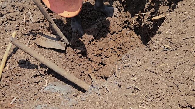 An orange pan is held in hand over parched, cracked earth after a worker digs the farm field with a pickaxe, a shovel resting nearby. It captures a harsh, labor-intensive scene of agriculture.