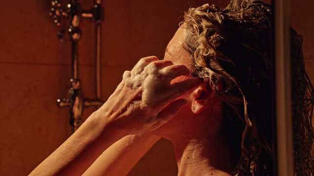 Young woman with long curly hair taking a shower in a dimly lit bathroom, gently massaging her scalp with shampoo for a relaxing and cleansing experience during her daily beauty routine