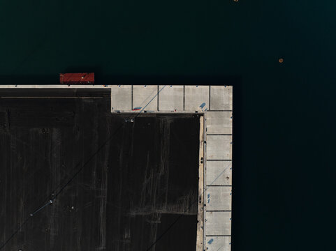 Aerial view of a geometric concrete pier corner meeting dark water with a small red container in Augusta, Sicily, Italy.