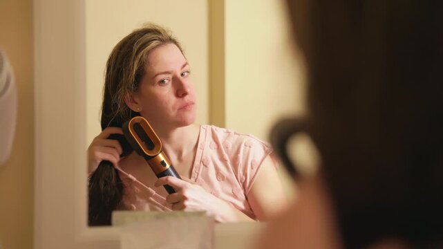 Beautiful woman using a modern hair dryer in front of a mirror during her daily routine. Personal grooming and self care concept showing her blow drying and styling her wet hair at home