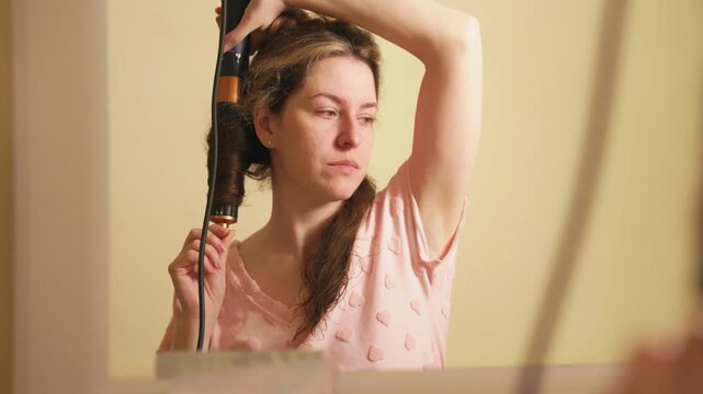 Millennial woman in pajamas styling her long brown hair with an automatic curling iron, looking at her reflection in the mirror as she prepares her hairstyle for a special event