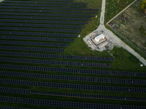 Aerial view of rows of solar panels in a photovoltaic power plant with a utility building and dirt path in Augusta, Sicily, Italy.