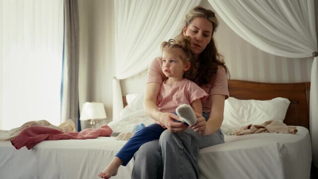 Young woman sitting on a bed carefully putting a sock on her little daughter's foot while getting her dressed for the day. Beautiful, caring moment between a loving parent and her child