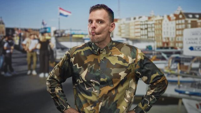 Man soldier in camouflage uniform with hands on hips and pursed lips, standing before canal buildings and boats on a street; confidence duty.