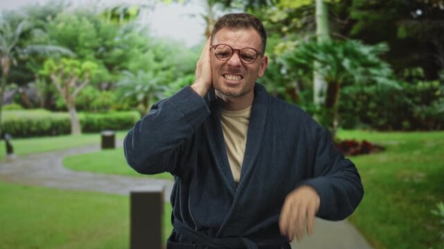 Man in navy robe and glasses holding his ear and grimacing while standing on a park walkway surrounded by trees in forest; earache distress.