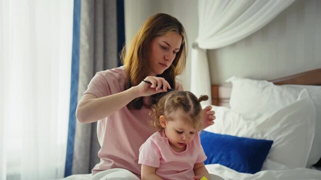 Loving mother sitting on a bed and carefully combing her little toddler daughter's curly hair, making a cute hairstyle for the child in a cozy bedroom during their morning routine