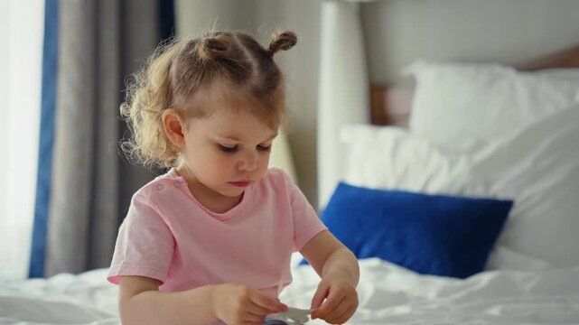 Adorable little girl with a cute hairstyle sitting on a comfortable bed, focused on a small object in her hands. A charming portrait of childhood curiosity, innocence, and peaceful play at home