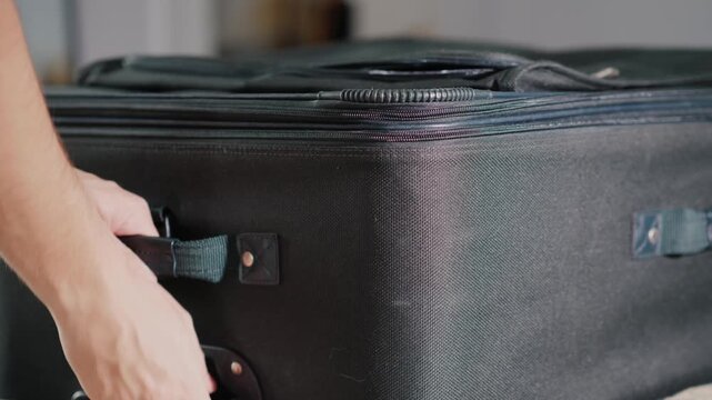 Hands closing large black fabric suitcase and taking it off bed in hotel room before traveling.