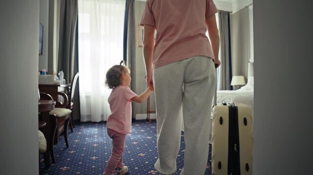 Young mother and her little daughter holding hands and entering a hotel room with a suitcase, ready to begin their family vacation and creating happy memories together on a trip
