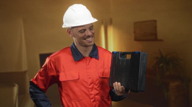 Man builder in hardhat holding a black toolbox and smiling while standing in a building livingroom with sofa and plant visible; calm confidence.