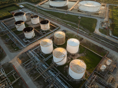 Aerial view of industrial oil storage tanks and complex pipeline networks surrounded by green fields during sunset in Augusta, Sicily, Italy.