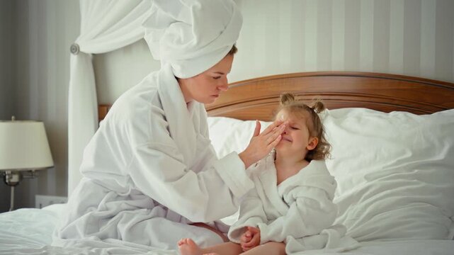 Young mother wearing a bathrobe and a towel on her head comforting her crying toddler daughter while sitting on a bed. Gentle care and motherhood concepts in a cozy home bedroom setting