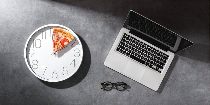 Pizza Time at work. White wall clock with pizza slice as hands, next to open laptop and black glasses on grey textured desk in soft sunlight . Creative lunch break concept. Top view, flat lay.