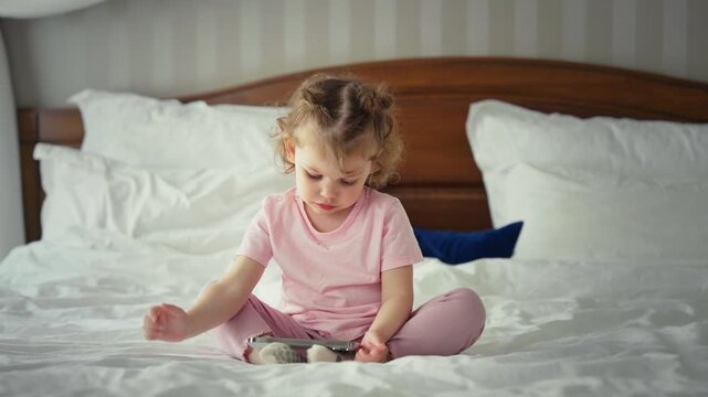 Adorable little girl with curly hair sitting on a big bed in her pajamas, completely absorbed in playing an educational game on a modern touchscreen smartphone before bedtime
