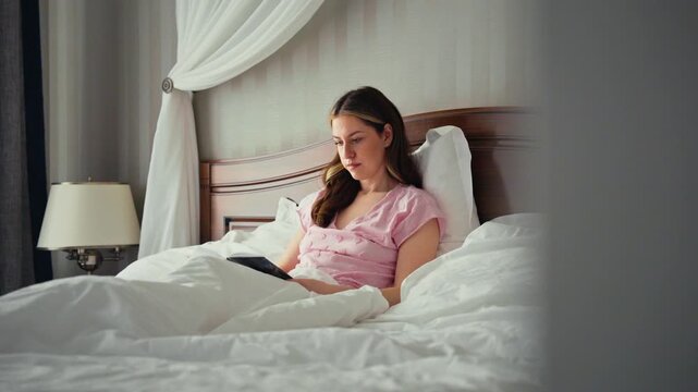 Young woman in pink pajamas relaxing and reading on an electronic reader while sitting in a comfortable bed with white linens, enjoying a quiet morning moment in a cozy bedroom at home