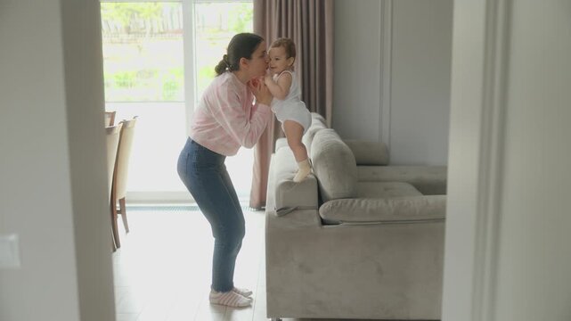 Mother helping baby girl stand on sofa backrest in bright living room