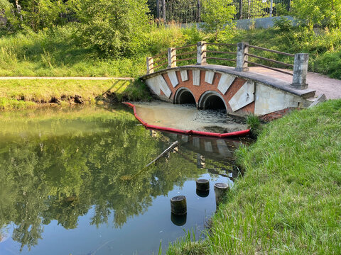 Arched pedestrian bridge over small canal with red oil boom protecting pond from pollution.