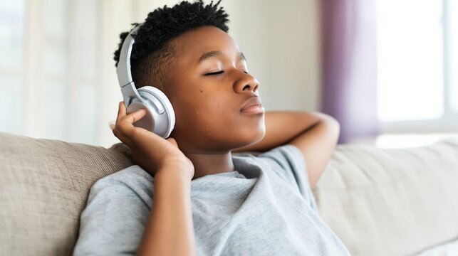 Boy relaxing on sofa with wireless headphones, eyes closed, listening to music at home.