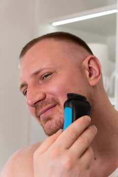 Vertical close up portrait of handsome young man shaving his face with electric shaver in bathroom mirror during morning routine. Male grooming, skincare and personal care concept.