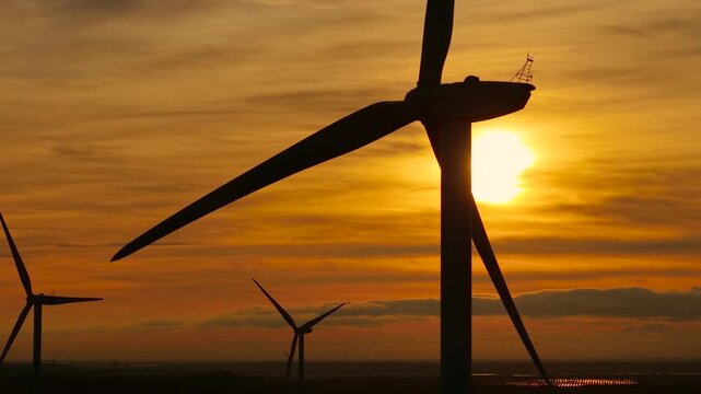 Spinning turbine backlit by sunset glow. Creating electricity with smooth wind. Producing renewable power from rotating blades. Delivering alternative energy using environmental force